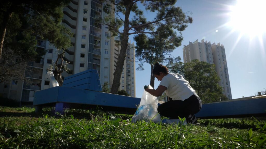 une femme ramasse des déchets sur la pelouse, on voit des immeubles derrière