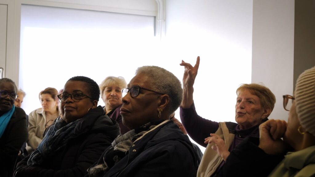 public de femmes d'un certain âge, dont quelques personnes racisées. une femme aux cheveux blancs lève la main